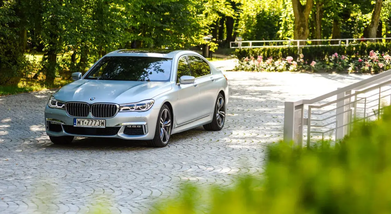 Srebrne BMW 750i lśni w słońcu na brukowanym podjeździe, otoczone zielenią i różami.