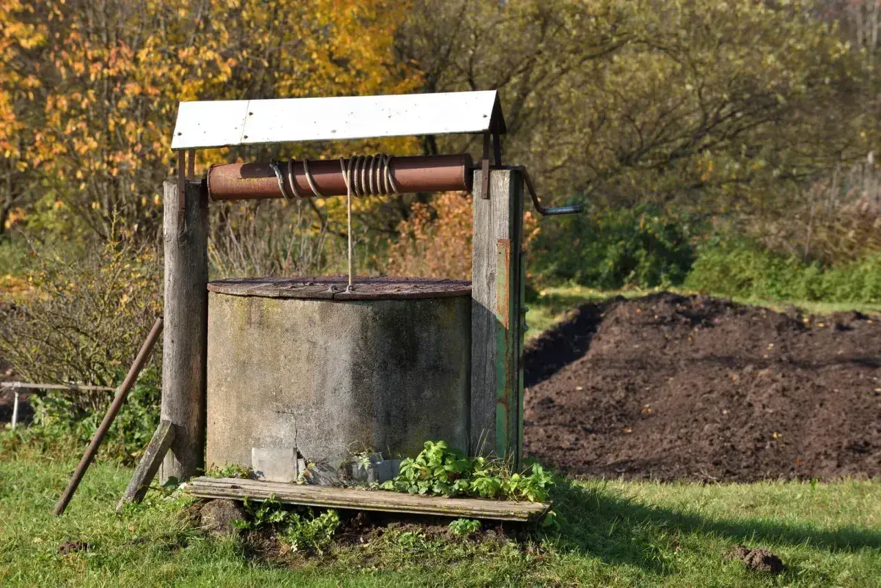 Stary, betonowy krąg studni kręgowej, z drewnianą konstrukcją i metalową korbą, otoczony jesienną zielenią.
