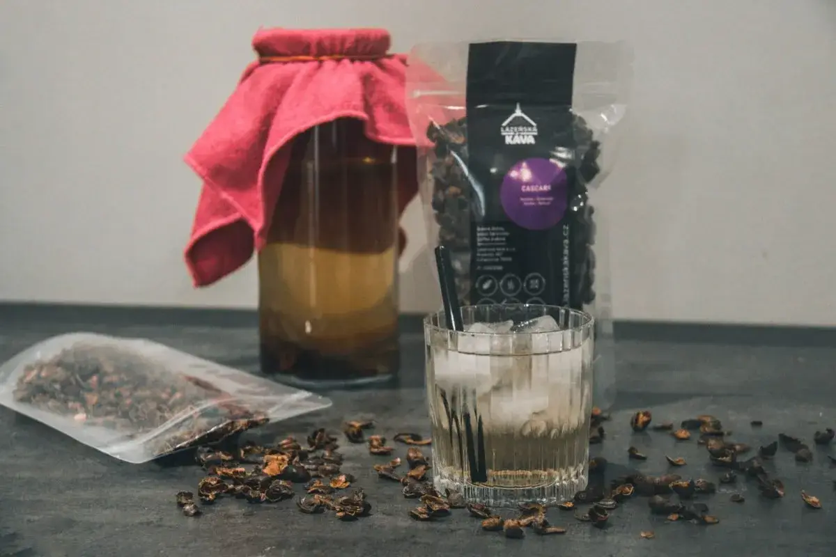 Napój z cascary, czyli co to jest kombucha z kawy, w szklance z lodem i słomką. Obok słoik z napojem i paczka z cascara.