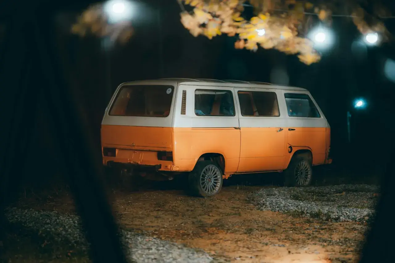 Pomarańczowo-biały kamper na nocnym parkingu, pod drzewami z lampkami. Czy można spać w kamperze na parkingu? Tak, to idealne miejsce na nocleg.