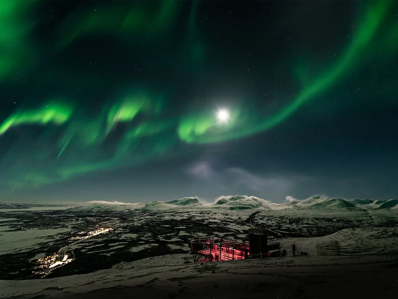 Norrsken över ett snöigt landskap med en upplyst byggnad. Kanske en vy från ett tåg till Abisko.
