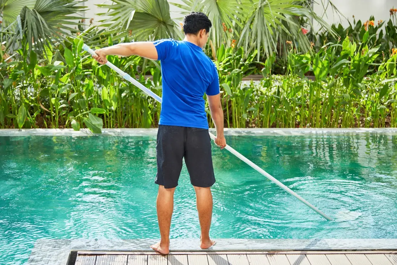 Hombre con camiseta azul y pantalones cortos negros limpia piscina de sal con un limpiafondos.
