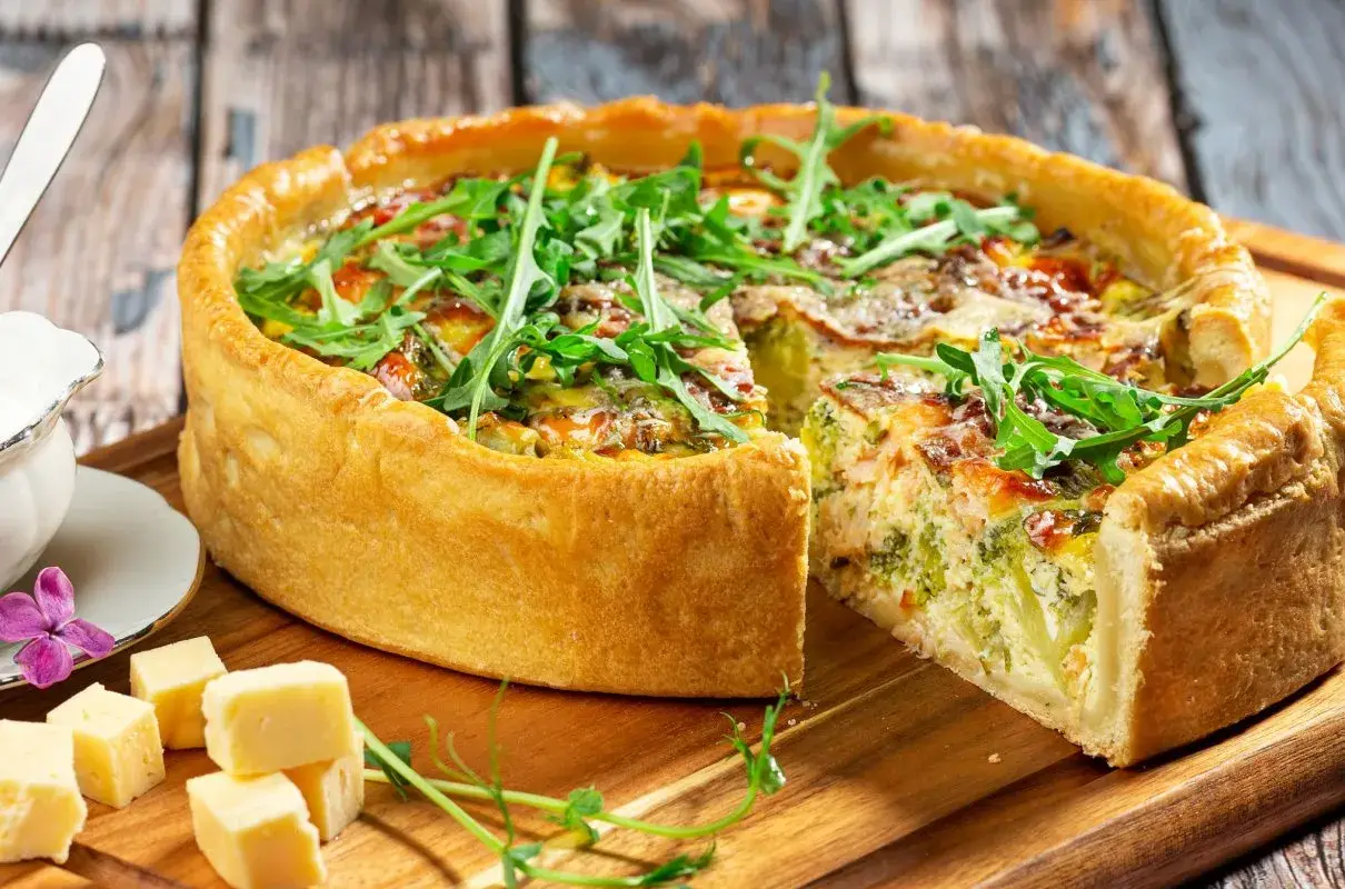 Pyszna tarta z brokułami i łososiem, ozdobiona rukolą. Obok kawałki sera i sos.