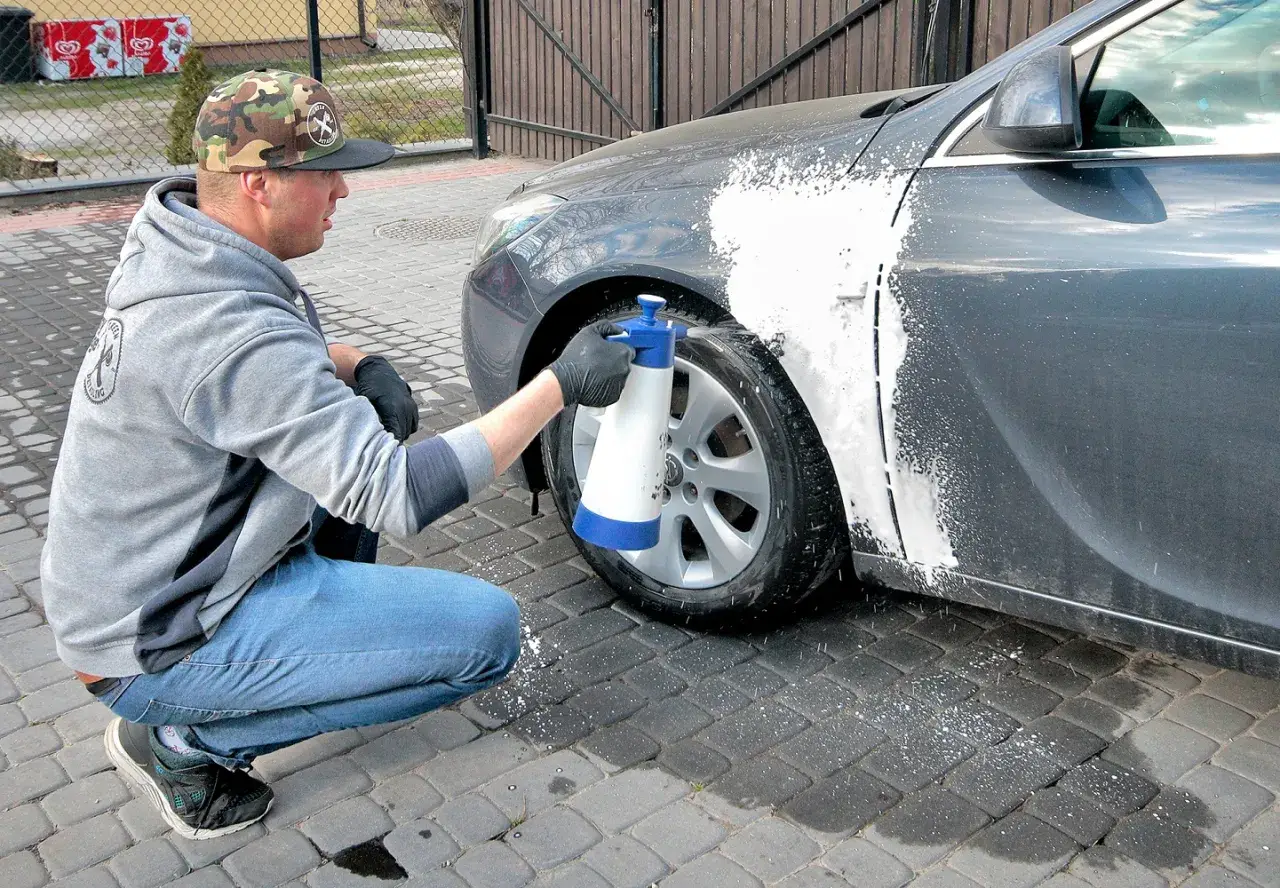 Mężczyzna w czapce z daszkiem i jeansach kuca przy samochodzie, rozpylając pianę na błotnik. Czy można naprawiać auto pod blokiem? Tak, jeśli to drobna naprawa lakieru.