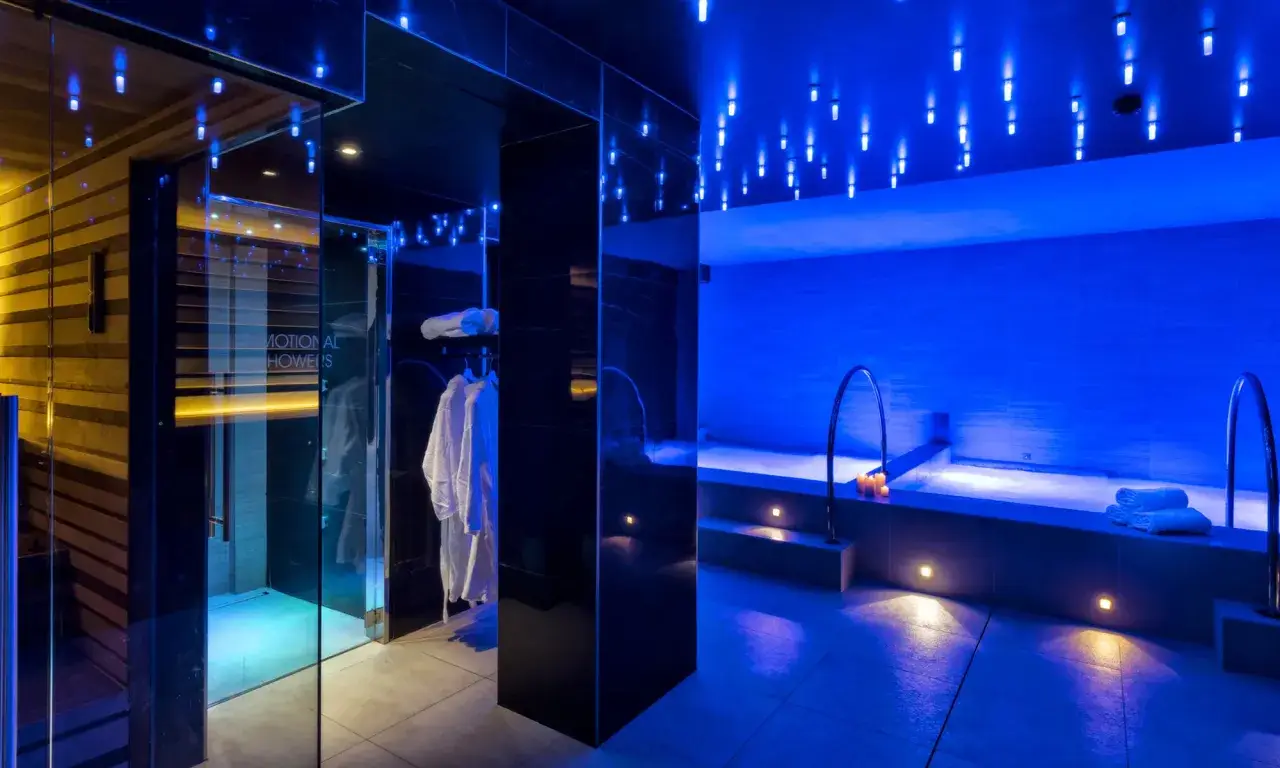 Interni di un centro benessere a Riccione con docce emozionali, vasca idromassaggio e accappatoi bianchi, illuminati da luci blu soffuse.