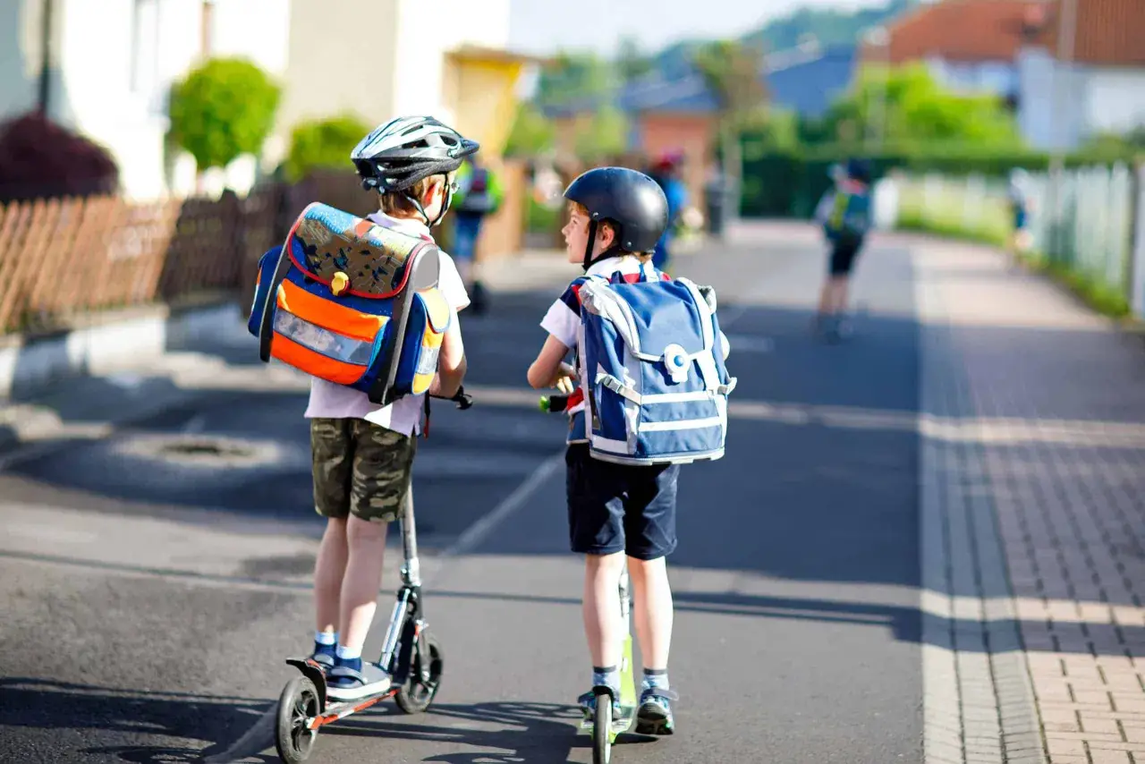 Deux enfants casqués, sacs à dos sur les épaules, se déplacent en trottinette. L'âge trottinette électrique est idéal pour ces jeunes écoliers.