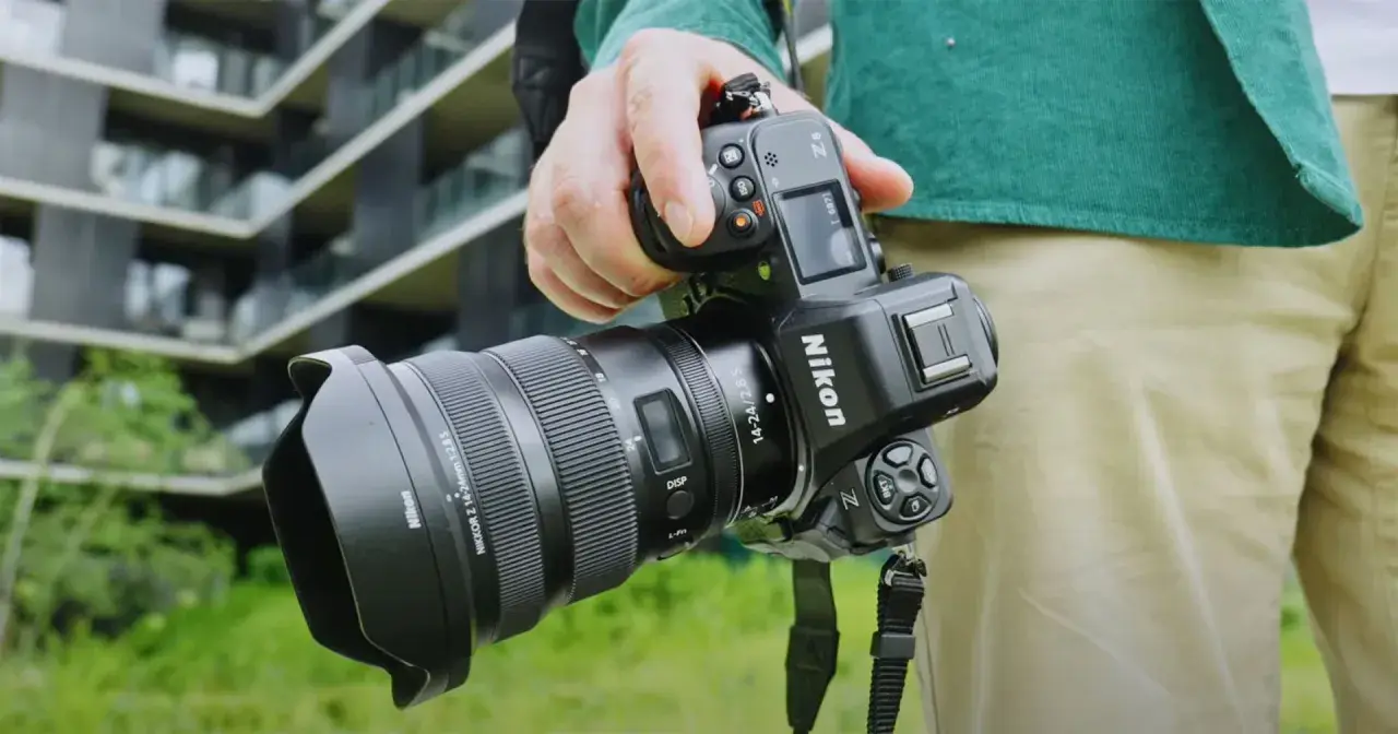Dłoń trzyma aparat Nikon Z8 z obiektywem. W tle nowoczesny budynek.