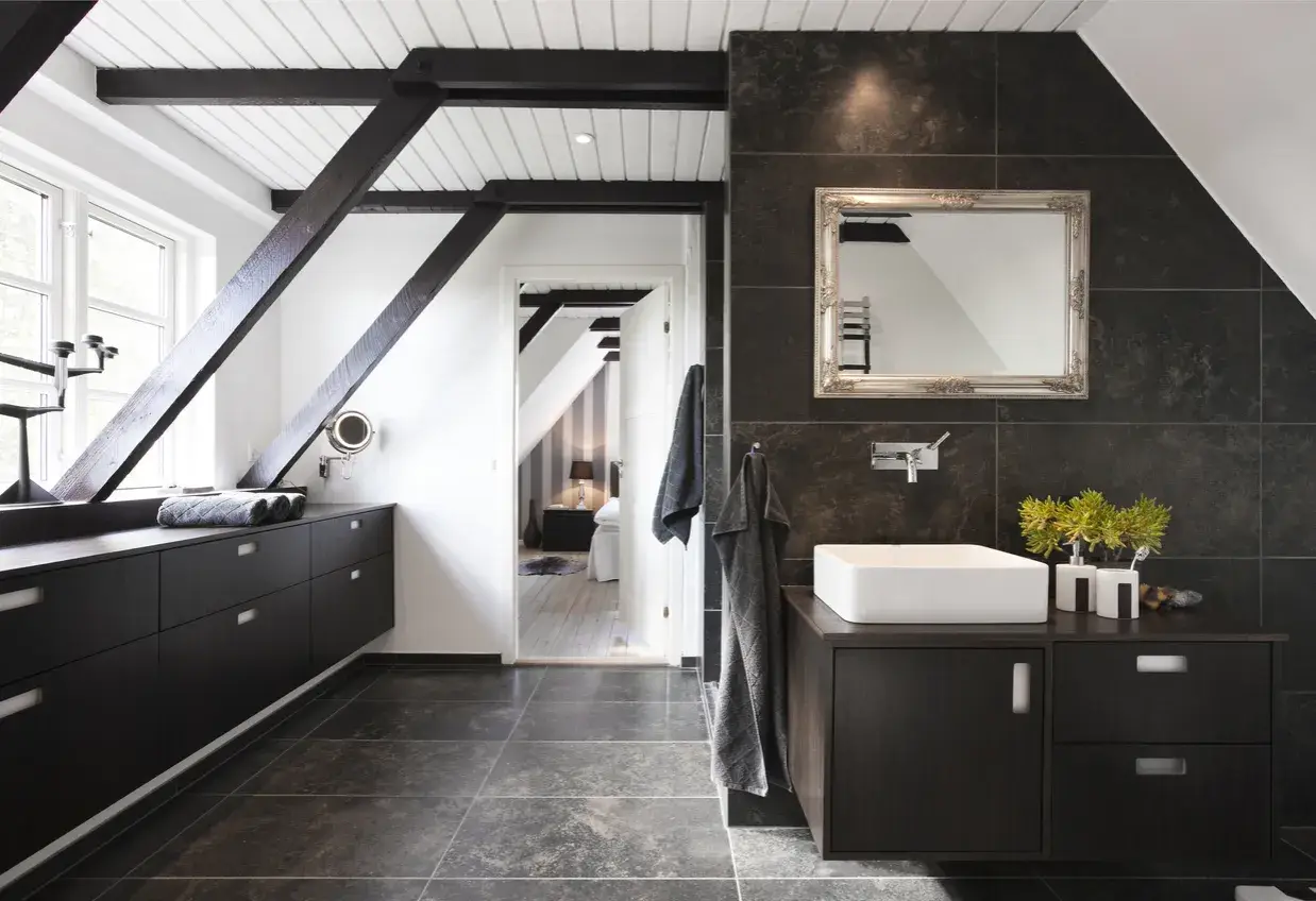 Baño moderno con vigas oscuras y suelo gris. El lavabo sobre encimera y los muebles oscuros contrastan con las paredes de baldosas oscuras. Se ve un dormitorio al fondo.