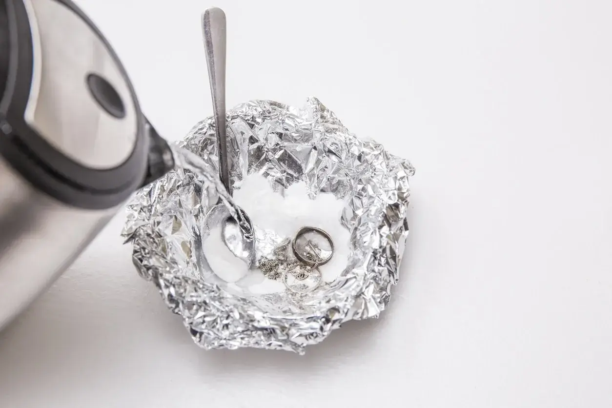 Nettoyage d'argenterie noircie : de l'eau chaude est versée sur des bijoux en argent et une cuillère dans une coupelle en papier aluminium.