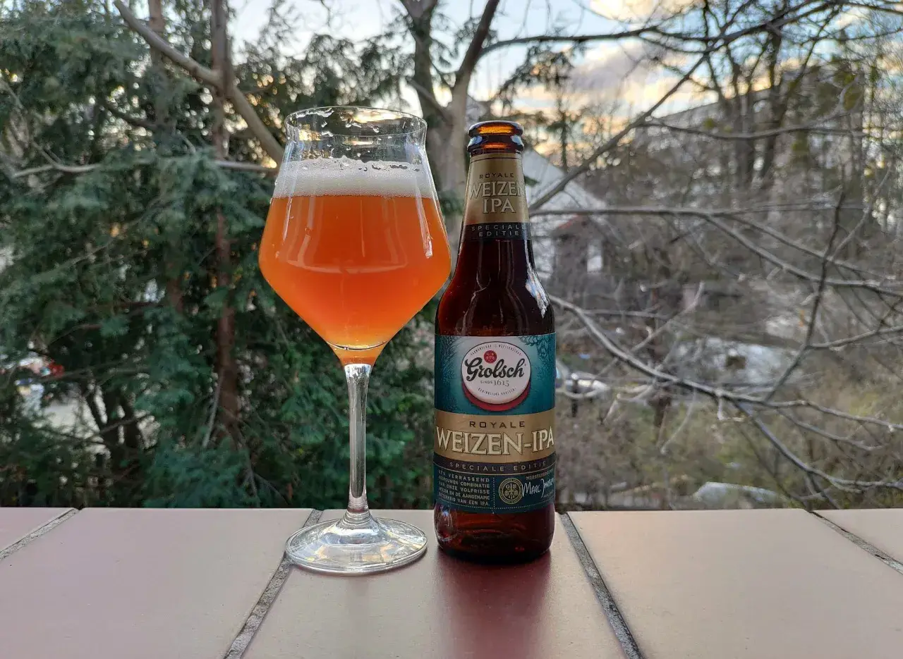 Szklanka z bursztynowym piwem Grolsch Royale Weizen IPA i butelka obok, na tle drzew i nieba.