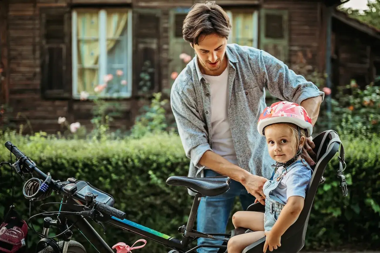 Tata zapina kask dziewczynce, która siedzi w foteliku rowerowym. Od kiedy można wozić dziecko na rowerze? Od kiedy jest bezpiecznie i ma kask!