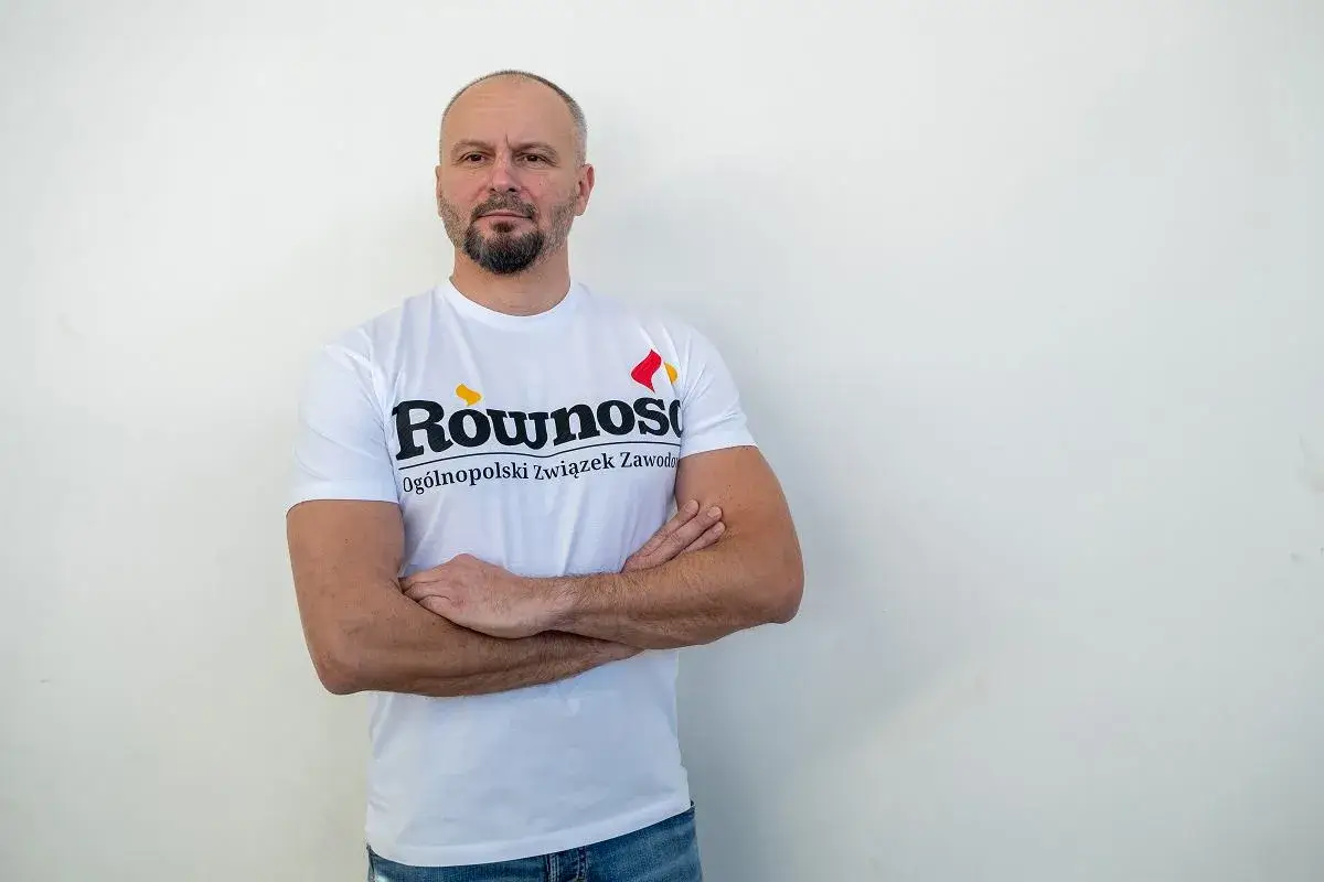 Mężczyzna w koszulce z napisem "Równość Ogólnopolski związek zawodowy" stoi z rękami skrzyżowanymi na piersi.