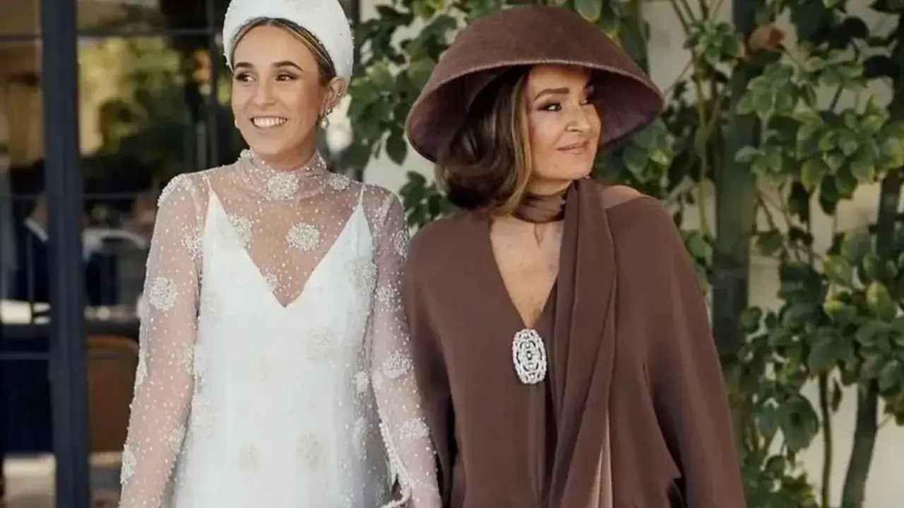 La madre de la novia, elegante con sombrero marrón, acompaña a la radiante novia con su vestido blanco adornado.