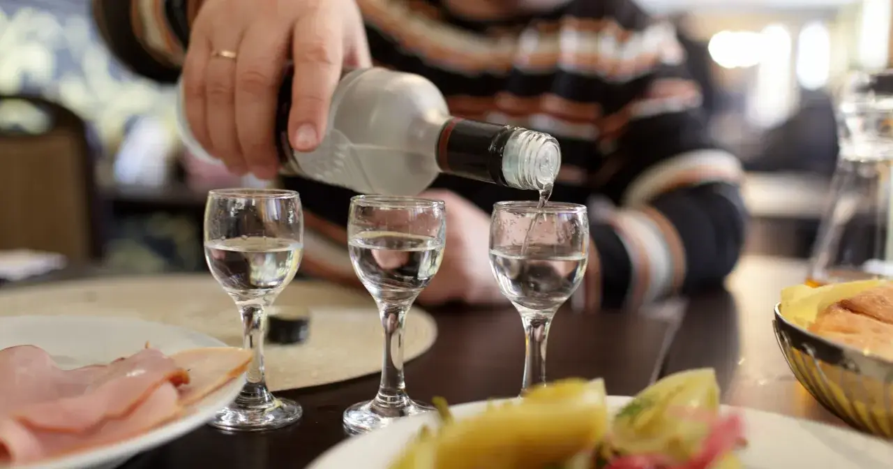 Ręka nalewa wódkę do kieliszków, przygotowując się na rekord promili. Na stole wędlina i przekąski.