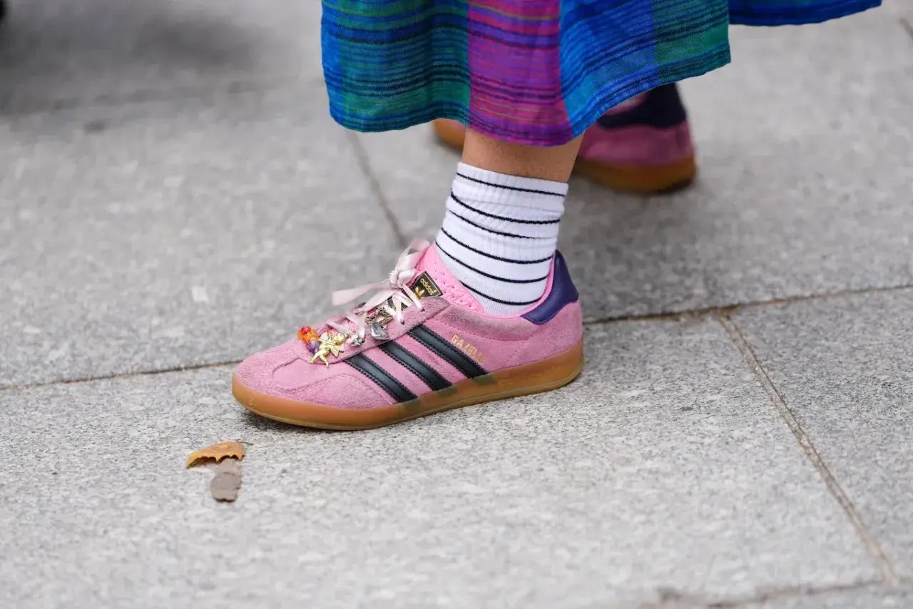 Różowe buty sportowe Adidas Gazelle z czarnymi paskami i charmsami, białe skarpetki w paski. W tle widać drugie, podobne buty w innym kolorze.