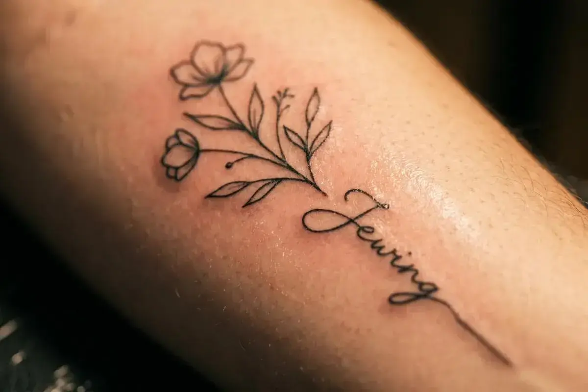 Delikatny line art tattoo z gałązką kwiatów i napisem "Sewing".