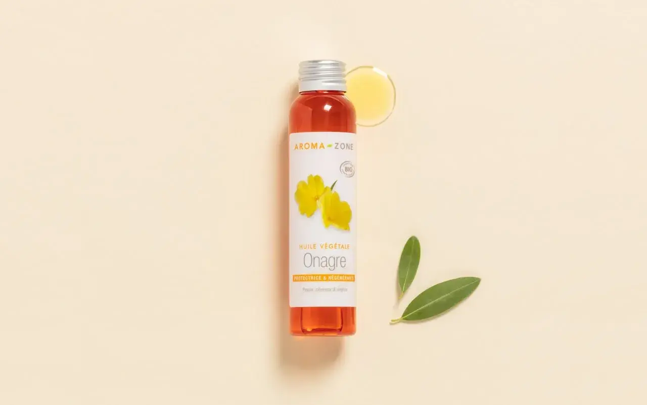 Flacon d'huile d'onagre Aroma-Zone, aux multiples bienfaits pour la peau et les cheveux. Une goutte d'huile dorée et deux feuilles vertes complètent la scène.