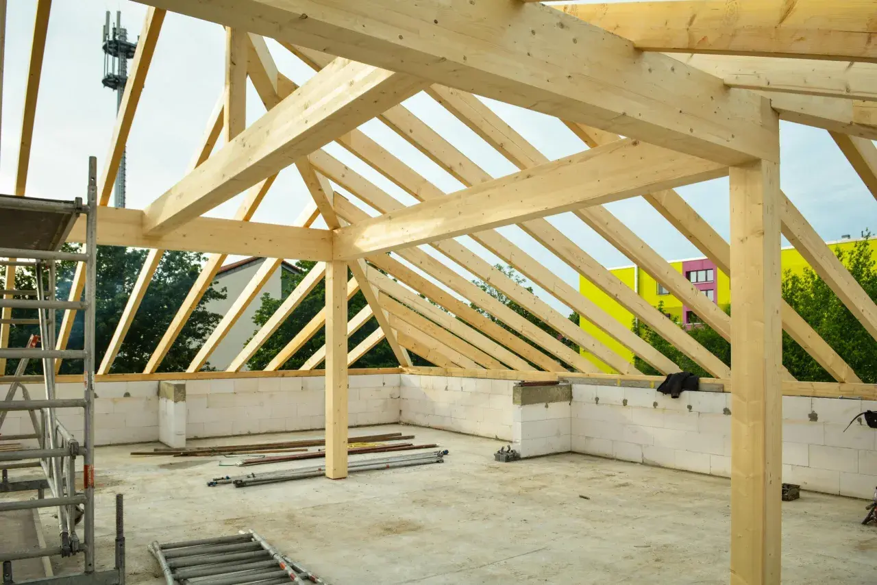 Struttura in legno per la realizzazione di tetti, con travi a vista e muri in blocchi di cemento. In sottofondo, attrezzature da cantiere.