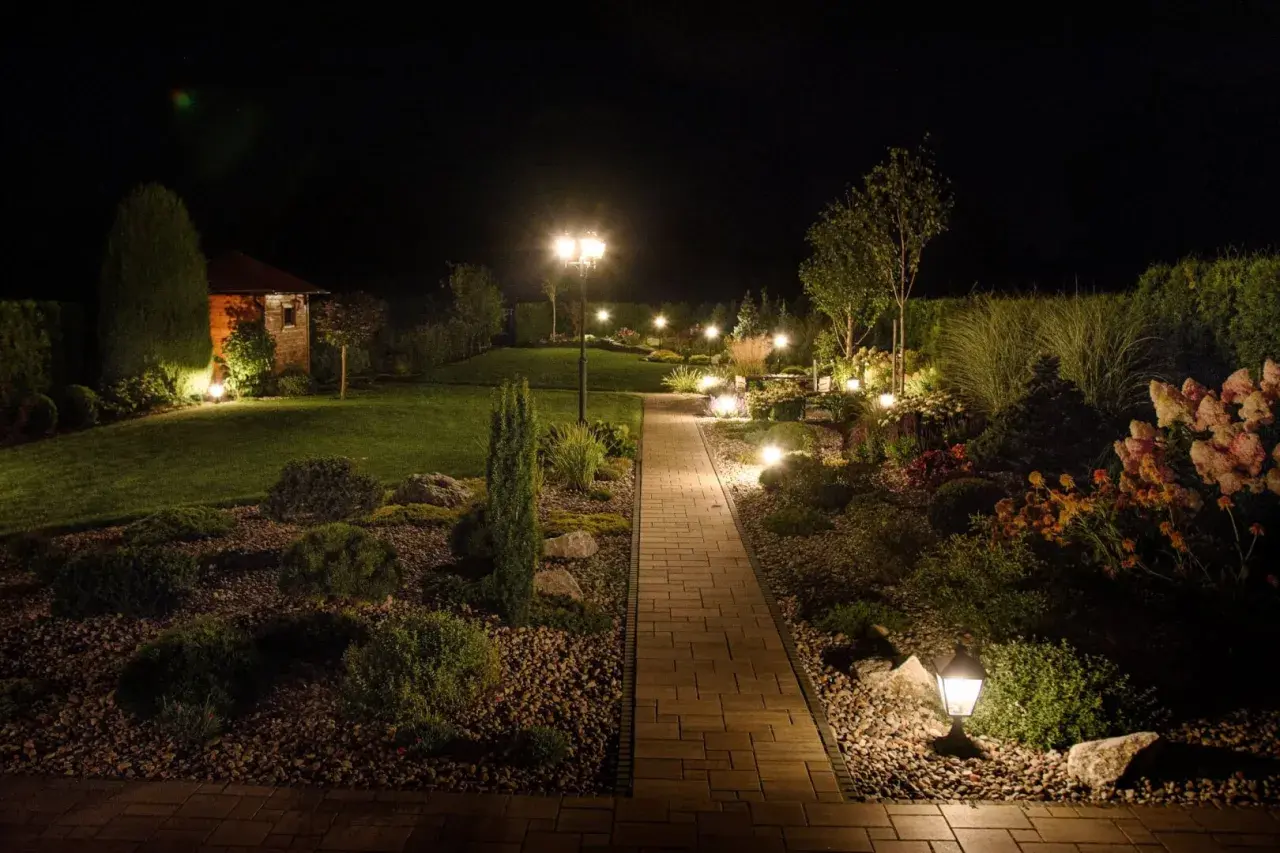 Piękna ścieżka z kostki brukowej prowadzi przez ogród, który nocą rozświetla nastrojowe oświetlenie. Inspiracje dla Twojego ogrodu.
