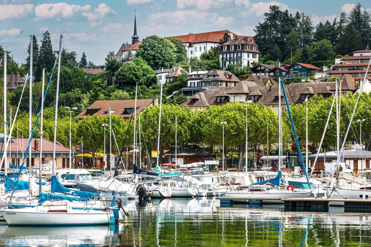 Port de plaisance à Thonon-les-Bains, avec des voiliers blancs et bleus amarrés, des maisons sur la colline et une église. Idéal pour savoir que faire à Thonon-les-Bains.