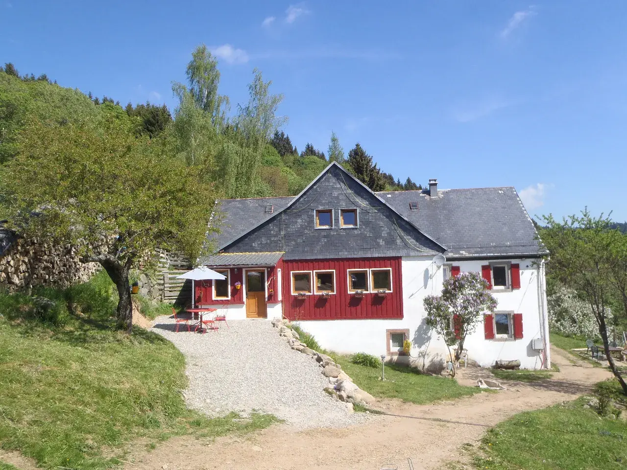 Maison de campagne avec terrasse et parasol, idéale pour un camping à la ferme dans les Vosges. Le soleil brille sur ce paysage verdoyant.