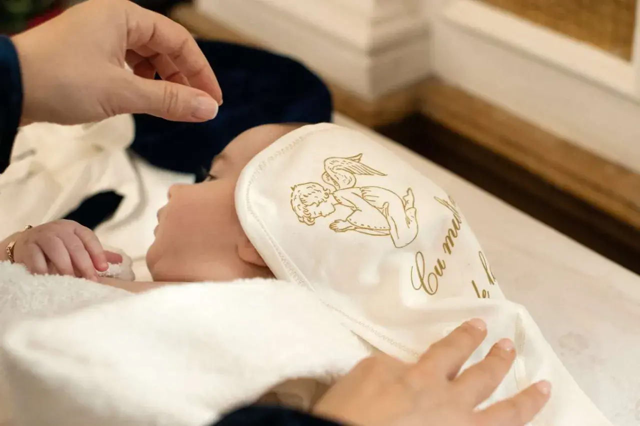 Dziecko otulone w biały kocyk z aniołkiem, przygotowywane do chrztu. To ważny moment, który potwierdzi zaświadczenie od księdza do chrztu.