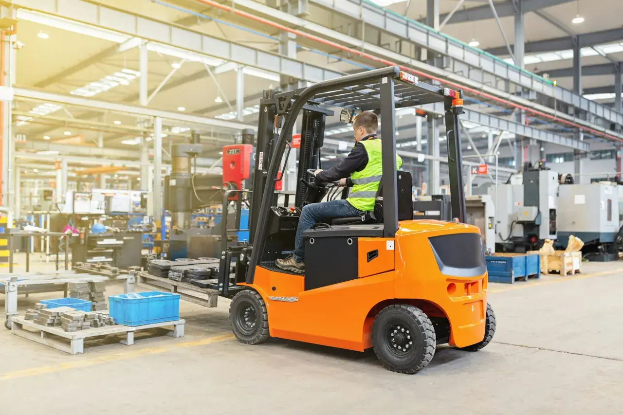 Ein Mann in Warnweste fährt einen orangefarbenen Gabelstapler in einer Fabrikhalle. Die Nutzungsdauer des Gabelstaplers ist entscheidend für die Effizienz.
