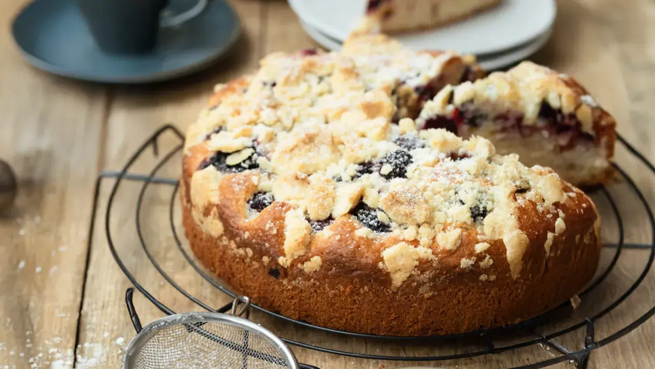 Ein saftiger, schneller Brombeerkuchen mit Streuseln, der auf einem Gitterrost abkühlt. Daneben eine Kaffeetasse und ein Stück Kuchen.