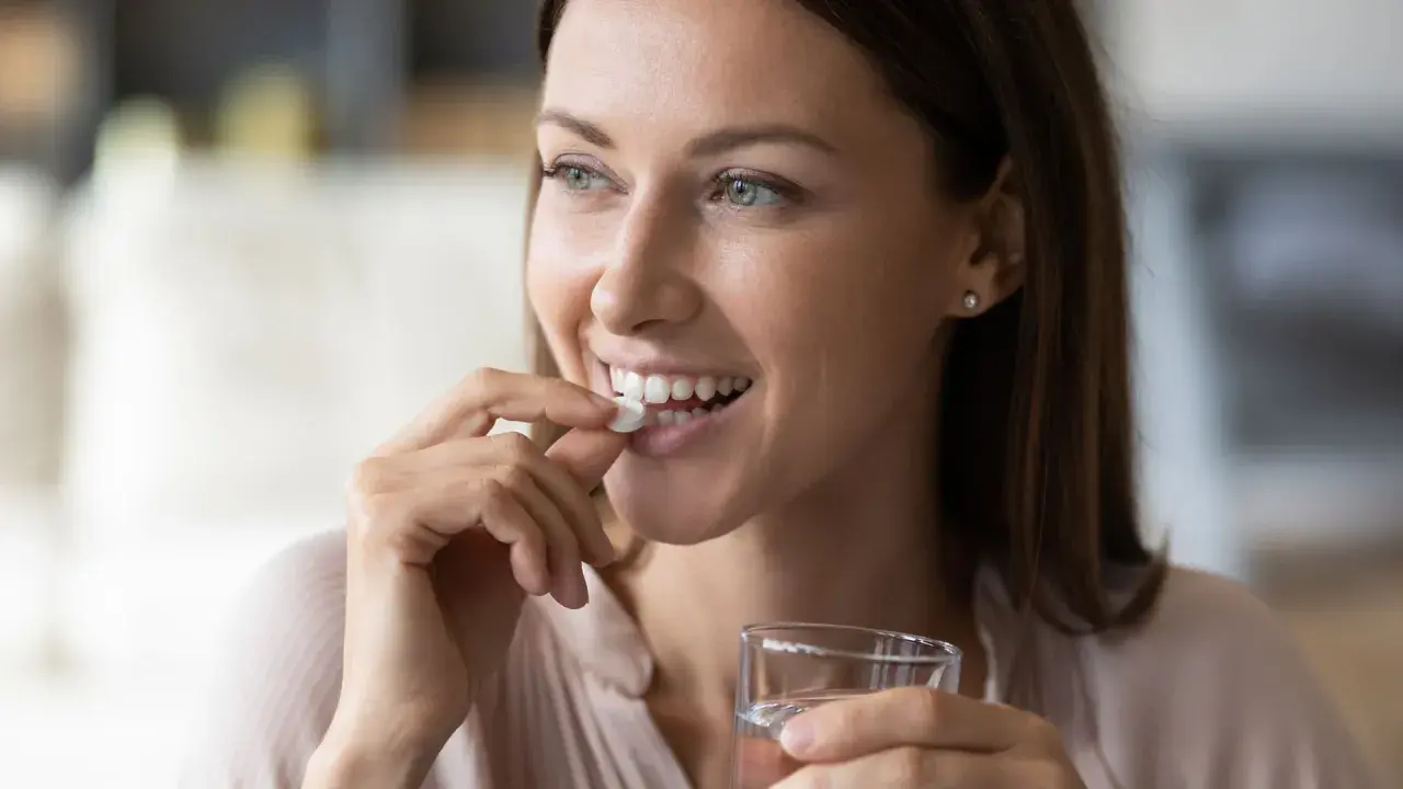 Femme souriante prenant un comprimé blanc, rappelant l'importance de ne pas associer le metronidazole alcool. Verre d'eau à la main.