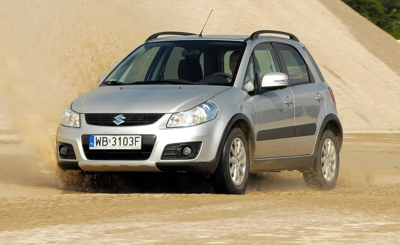 Srebrny Suzuki SX4 pokonuje piaszczystą wydmę, wzbijając tumany kurzu.