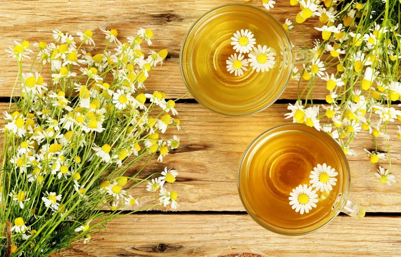 Due tazze di tisana dorata con fiori di camomilla galleggianti, accanto a un mazzo di fiori freschi. Rimedio naturale per il bruciore di stomaco.