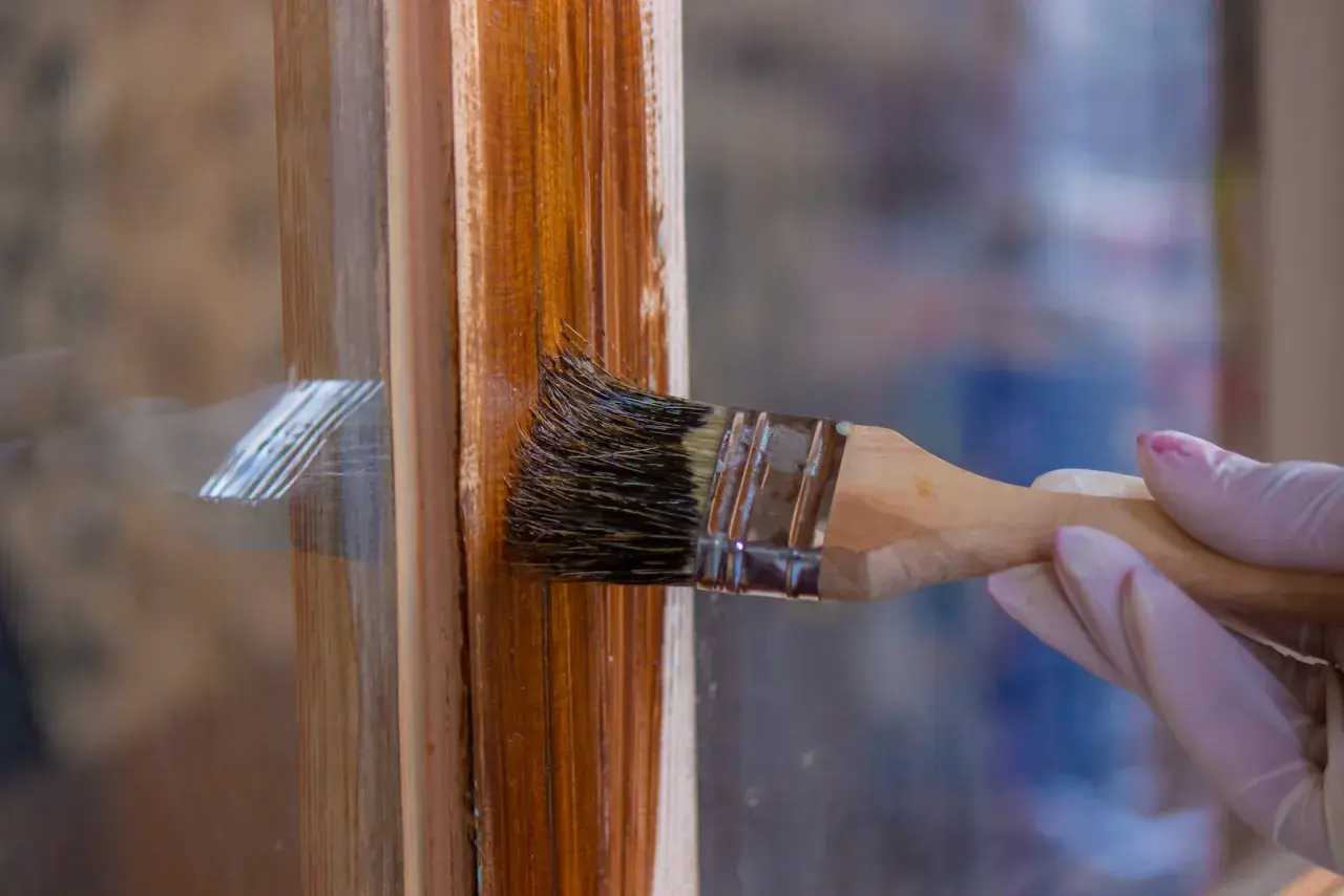 Eine Hand mit Handschuh streicht mit einem Pinsel einen braunen Holzrahmen. Das Holzfenster erstrahlt bald in neuem Glanz.