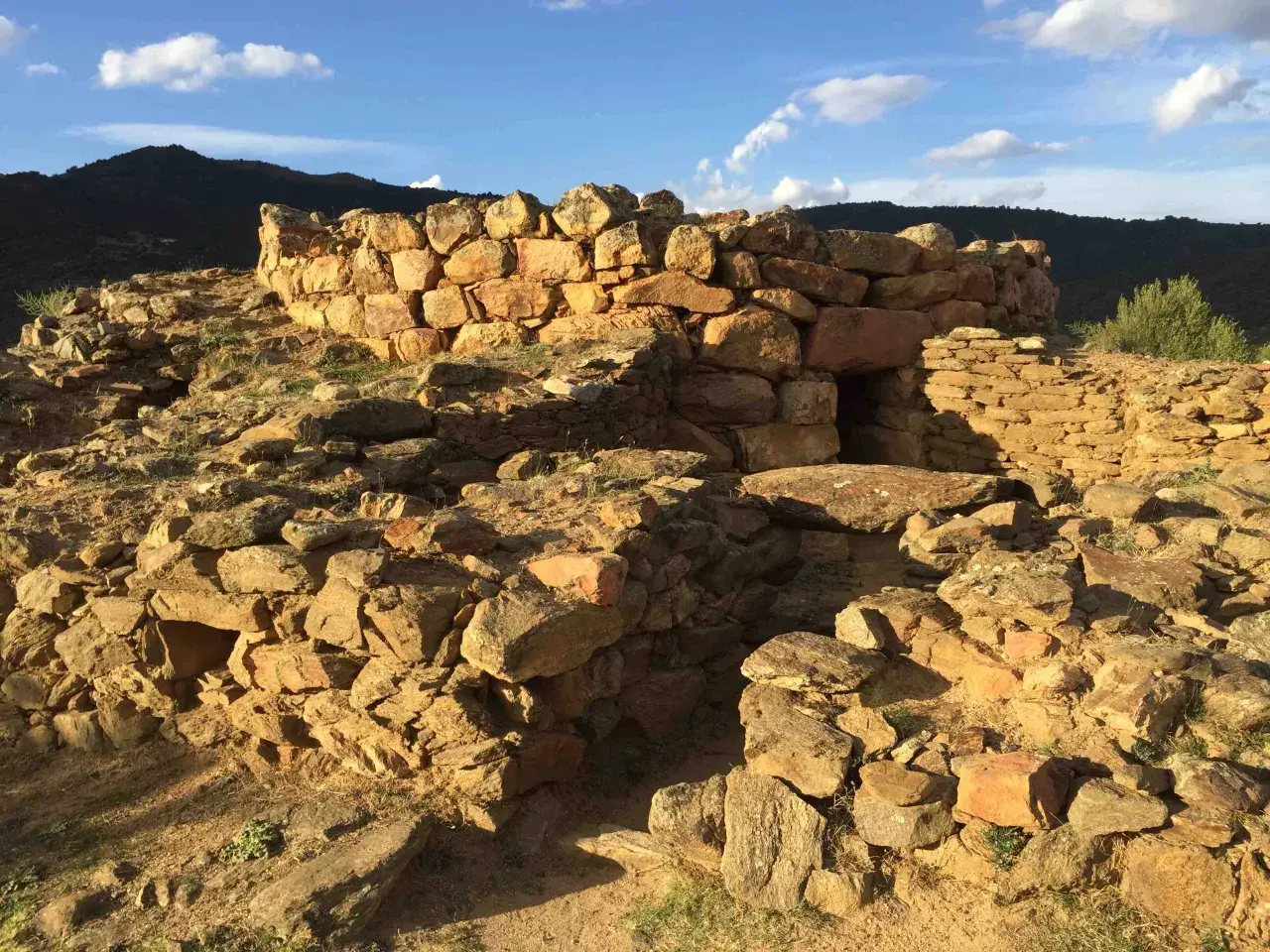 Rovine del nuraghe San Pietro, una struttura in pietra con un ingresso scuro, sotto un cielo azzurro con nuvole bianche.