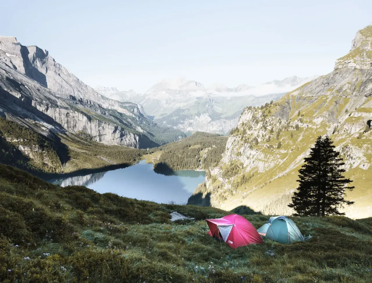 Two tents are set up on a grassy slope overlooking a serene lake in the Swiss Alps. This is a perfect spot for swiss alps hiking and camping.