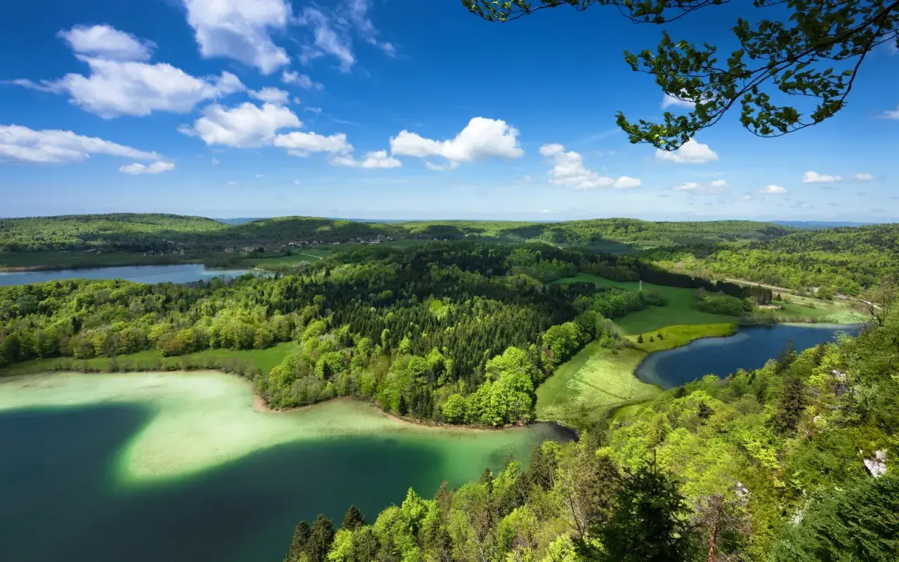 Paysage verdoyant du Jura avec des lacs aux eaux claires, des forêts denses et un ciel bleu parsemé de nuages blancs.