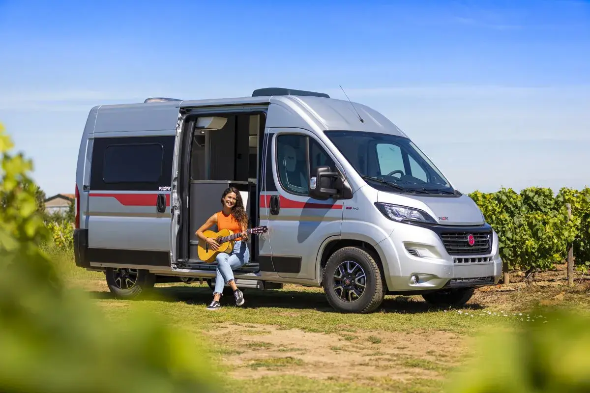 Une jeune femme joue de la guitare assise à l'entrée d'un camping-car aménagé, un type de camping car idéal pour les escapades ensoleillées.