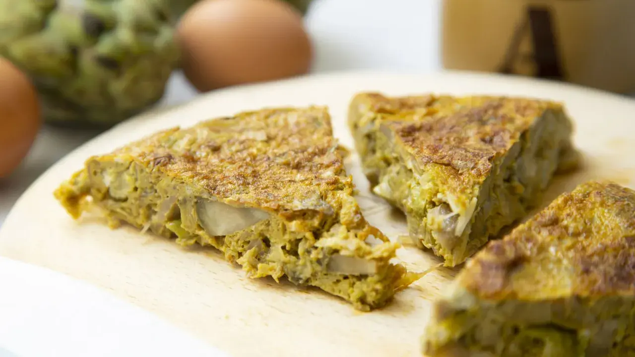 Fette di frittata con pecorino e verdure su tagliere di legno, con uova e carciofi sullo sfondo.