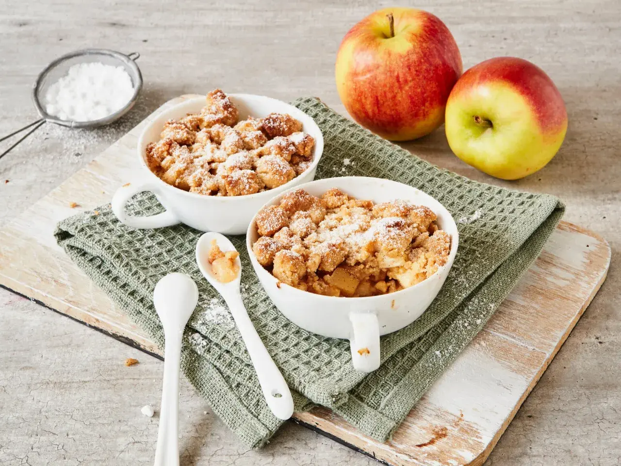 Zwei Schalen mit warmem Apfel Crumble, bestäubt mit Puderzucker. Daneben liegen zwei Äpfel und Löffel.