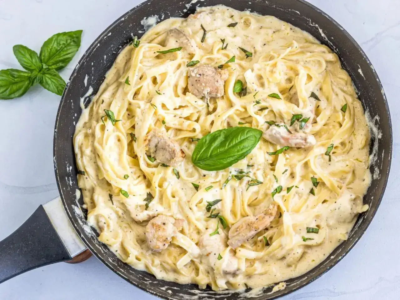 Un cremoso plato de pasta alfredo con trozos de pollo y hojas de albahaca fresca en una sartén.