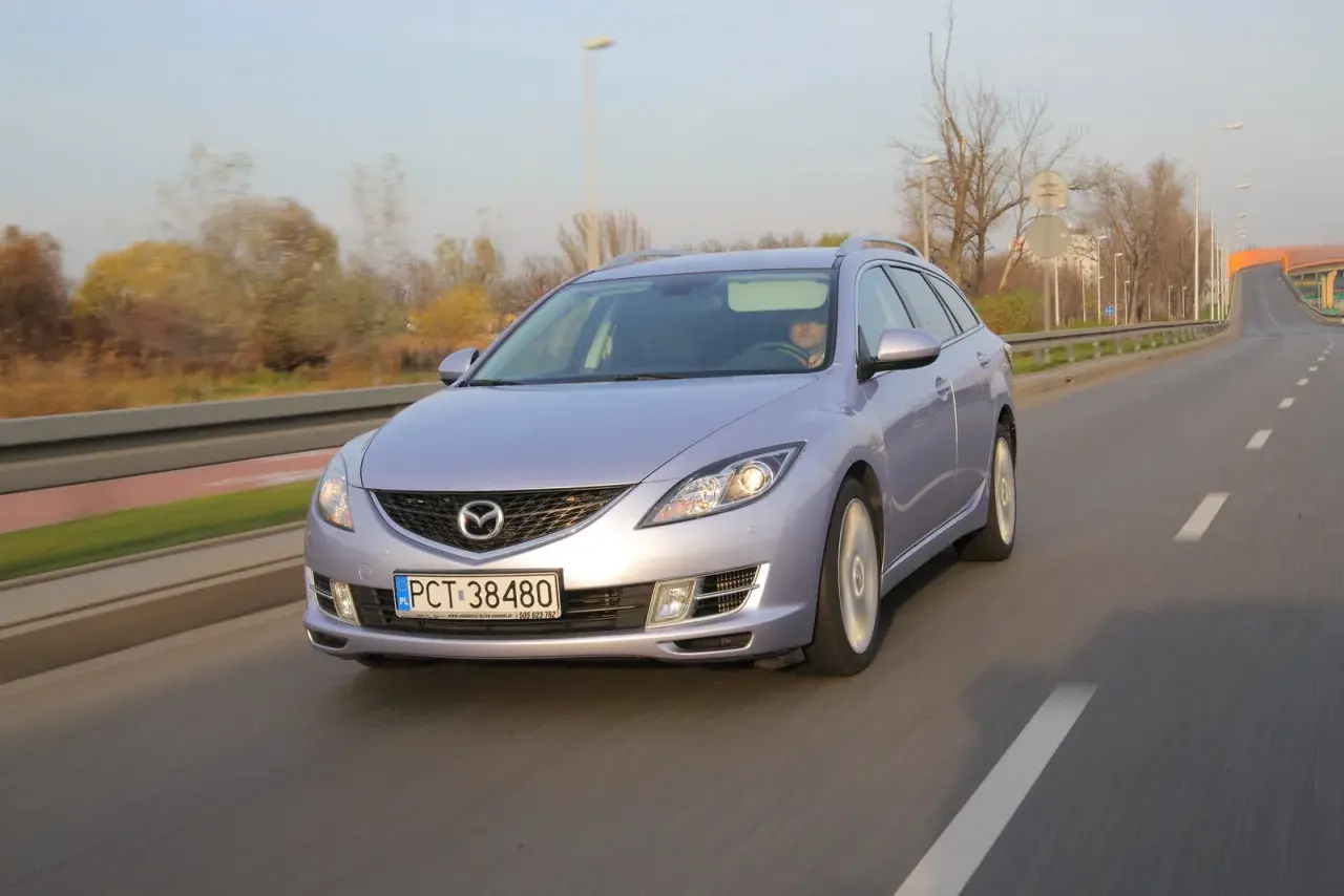Fioletowa Mazda 6 GH jedzie po drodze. Jej żarówki świecą jasno, oświetlając trasę.