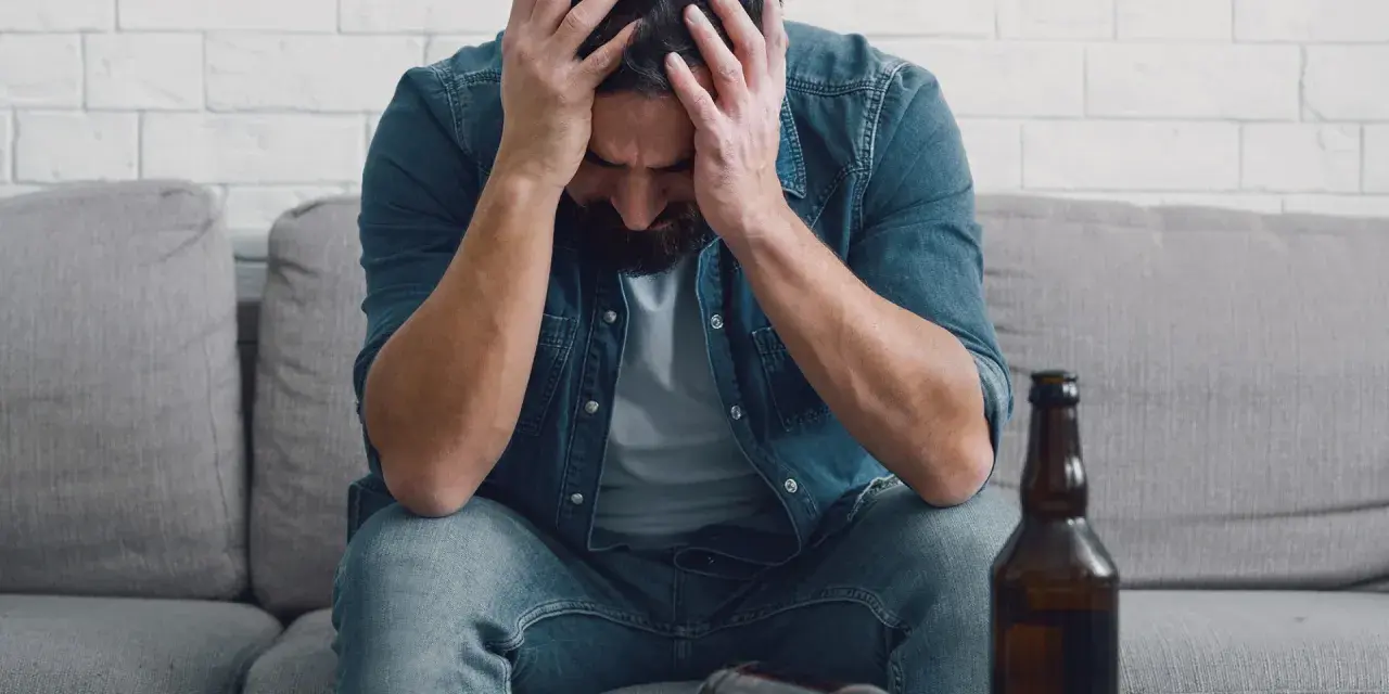 Man met handen in het haar, zittend op een bank met een fles bier. Dit illustreert de definitie van verslaving: een dwangmatige behoefte.