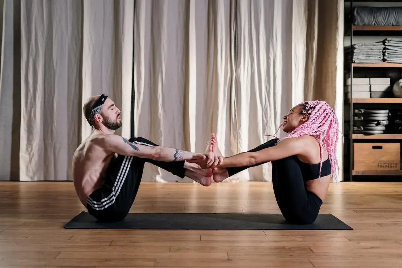 Un homme et une femme, débutants en yoga, pratiquent une posture à deux. Ils se tiennent les mains et les pieds, créant une connexion harmonieuse.