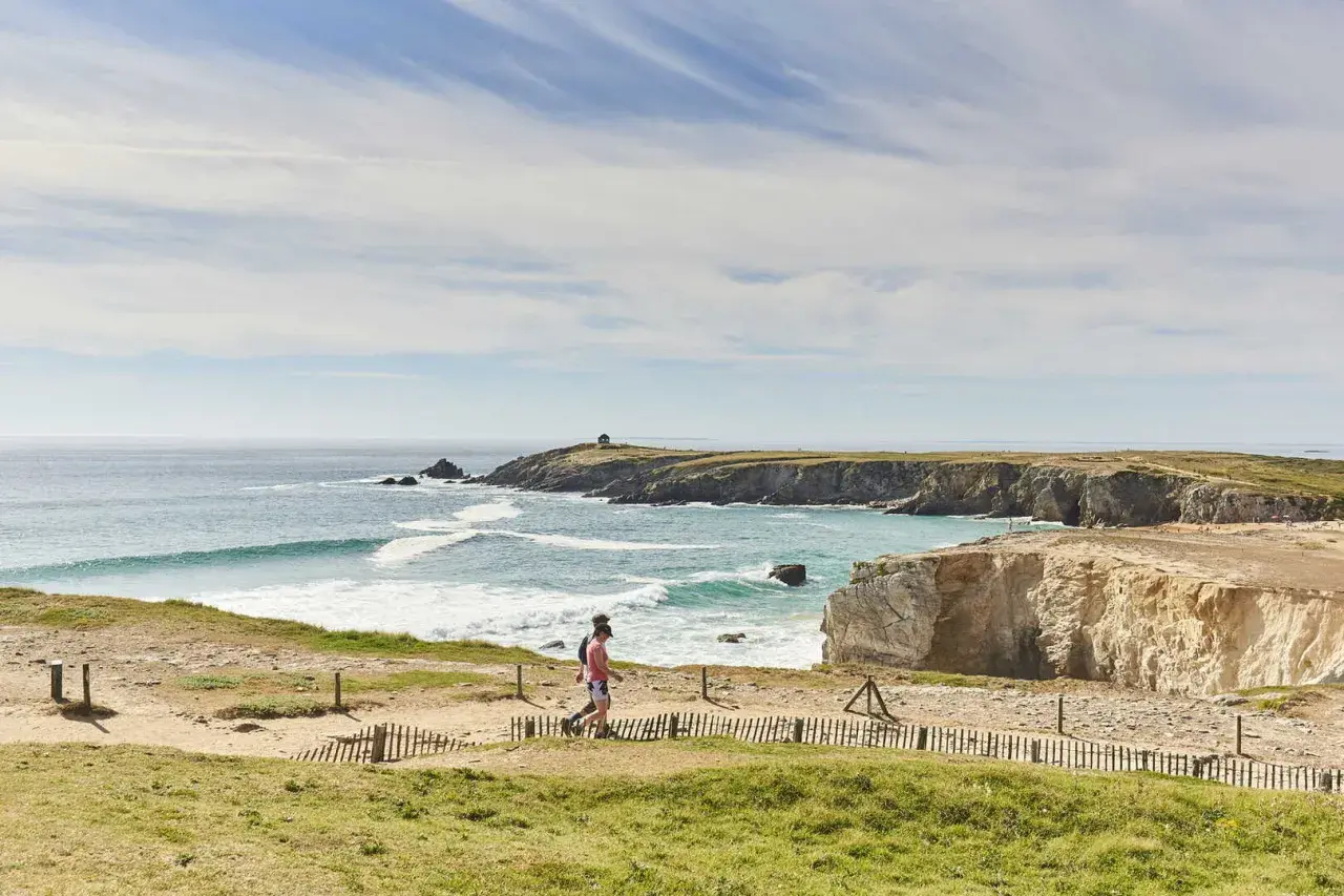 Deux randonneurs profitent du tour de la presqu'île de Quiberon à pied, longeant la côte rocheuse et la mer agitée sous un ciel bleu.