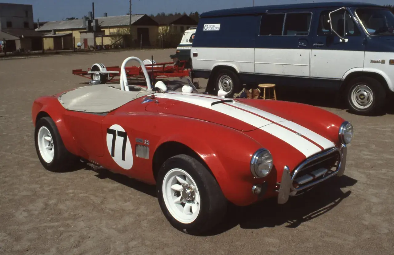 Röd sportbil, en kungens AC Cobra med nummer 77, redo för race. Bakom skymtar en blåvit skåpbil.