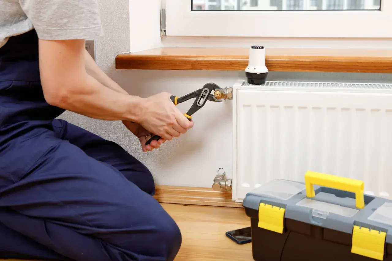 Instalator dokręca kluczem złączkę S do grzejnika, przygotowując go do sezonu grzewczego. Obok stoi skrzynka z narzędziami.