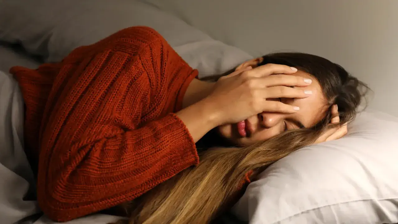 Mujer con suéter naranja se tapa la cara, agotada. Dormir poco engorda y afecta tu bienestar.