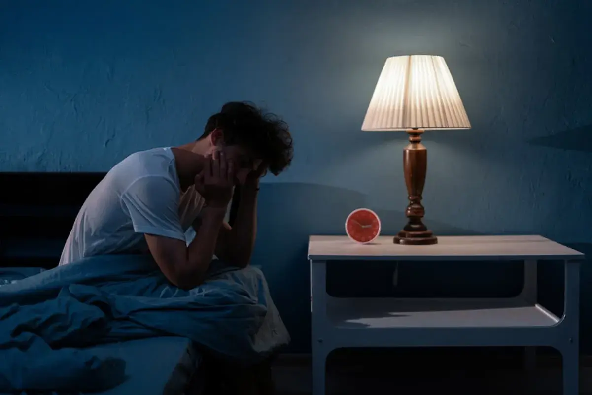 Un joven sentado en la cama, con la cabeza entre las manos, ilustra qué pasa si no duermes una noche. La lámpara de noche y el reloj marcan la hora.