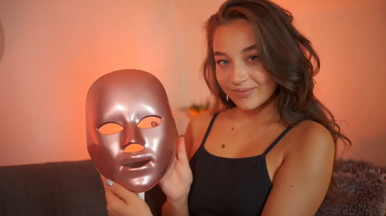 En ung kvinna håller upp en roséguld LED-mask för ansiktet, redo för en led-ljus behandling ansikte.