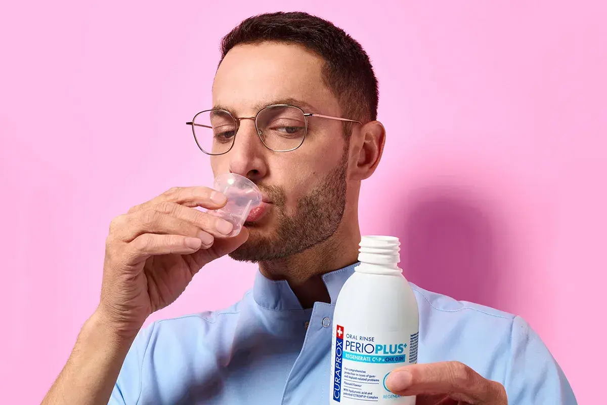 Uomo con occhiali beve collutorio da un bicchierino. Il flacone di Curasept gel va risciacquato.