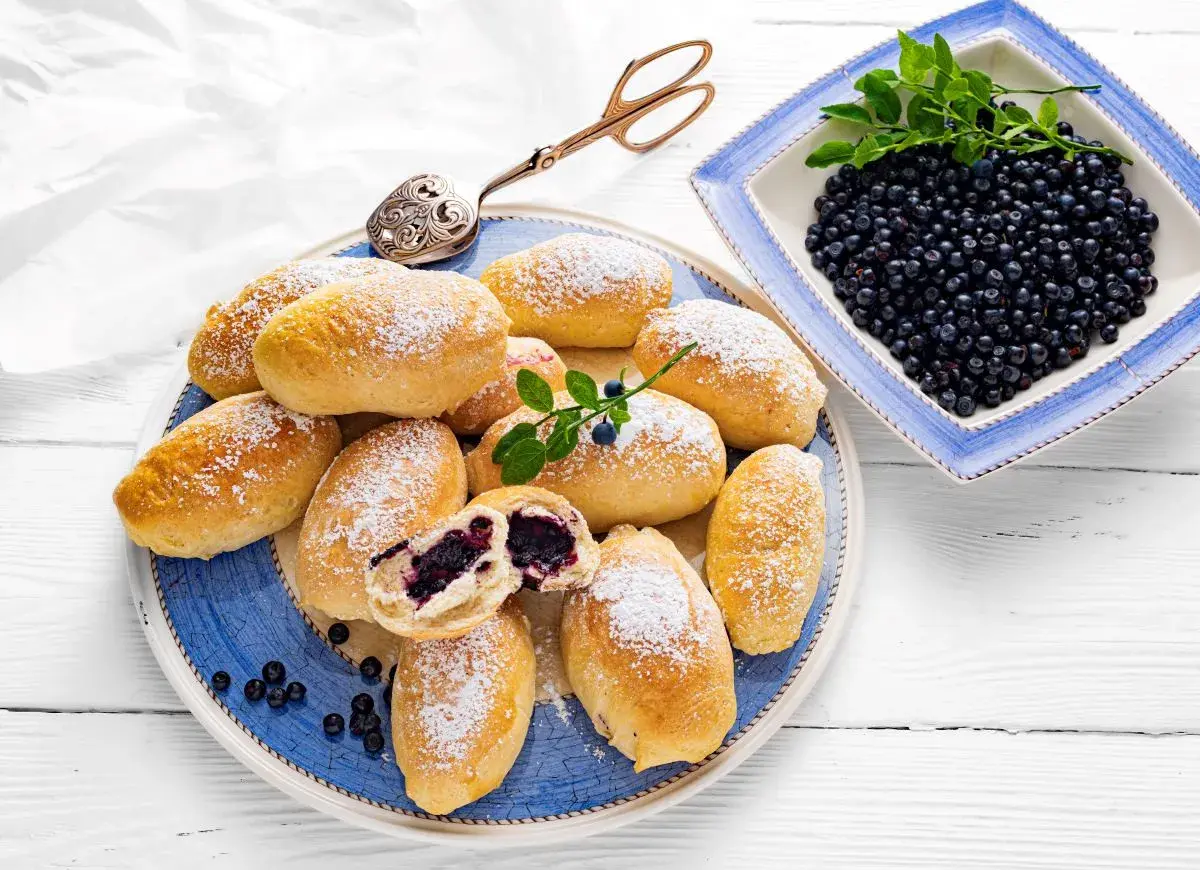 Pyszne jagodzianki posypane cukrem pudrem, obok miseczka świeżych jagód. Idealne na lato, zgodnie z najlepszym jagodzianki przepis.